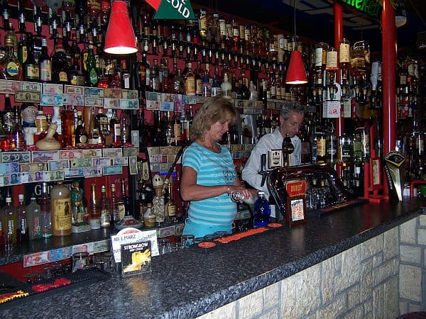 Der Tresen der 08/15 Bar mit Zapfanlage, roten Pendelleuchten und zwei Personen hinter der Theke.